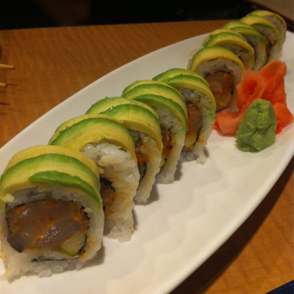 Sushi roll topped with avocado slices on a white rectangular plate, accompanied by pickled ginger and wasabi.
