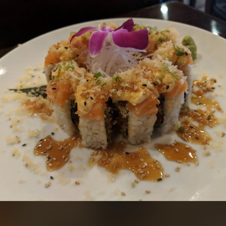 Sushi rolls topped with crispy crumbs, salmon, sesame seeds, sauces, shredded daikon, and a purple orchid garnish on a white plate.