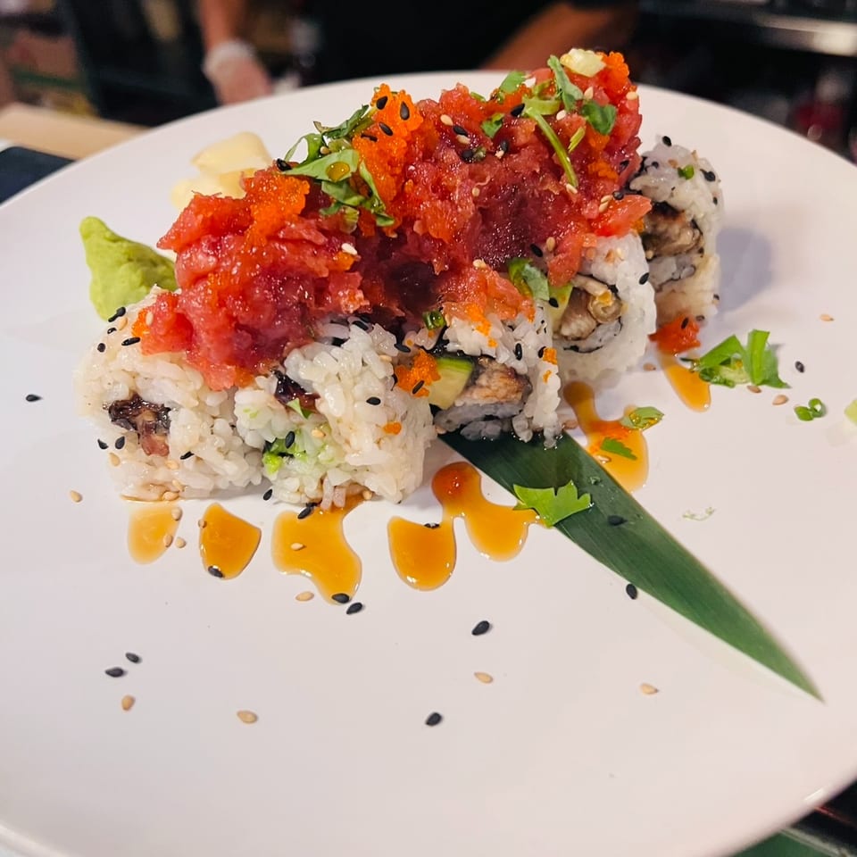 Sushi roll topped with spicy tuna, orange roe, greens, sesame seeds, and drizzled sauce on a white plate.