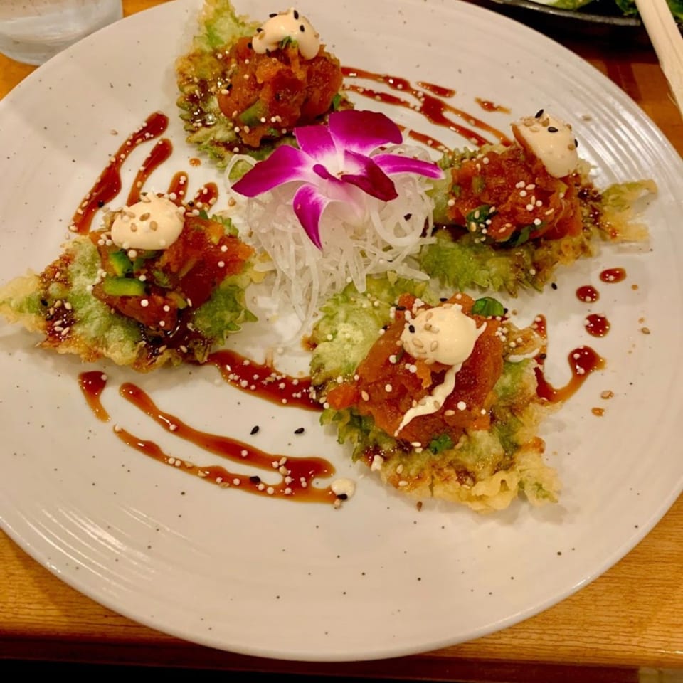 Platter of four crispy sushi-style bites with spicy topping, creamy sauce, sesame seeds, lettuce, and a purple orchid garnish.