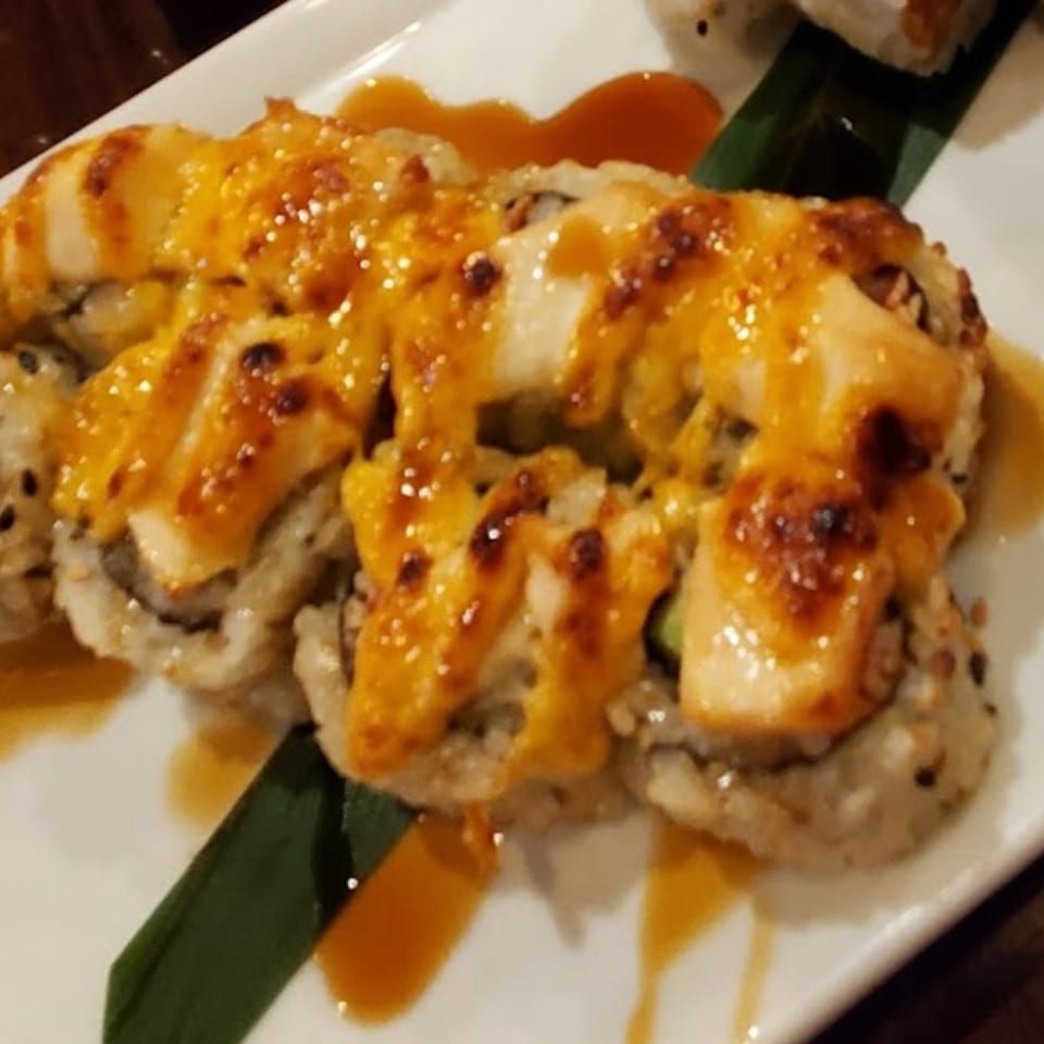 Sushi rolls topped with grilled salmon, creamy orange sauce, and sweet glaze on a white plate with a green leaf garnish.