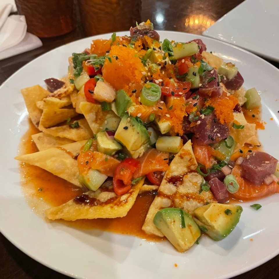Loaded nachos topped with diced avocado, tomatoes, onions, sesame seeds, orange roe, and cubed raw fish on a white plate.