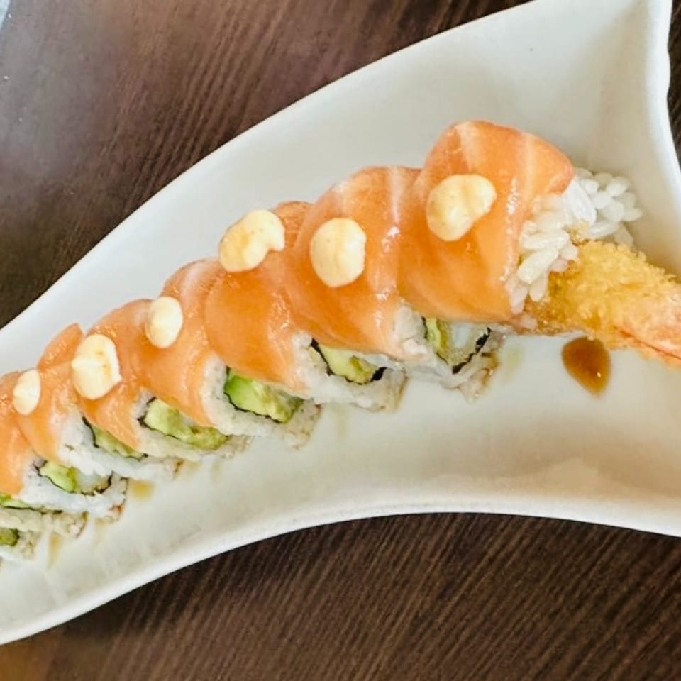 Salmon-topped sushi roll with avocado, creamy sauce, and tempura shrimp served on a white curved plate.
