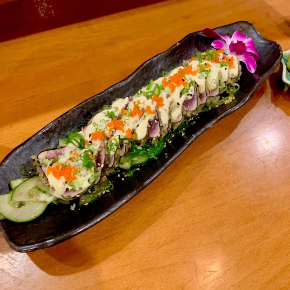 Sushi roll topped with creamy sauce, orange roe, scallions, and sesame seeds on a black platter with cucumber and orchid garnish.