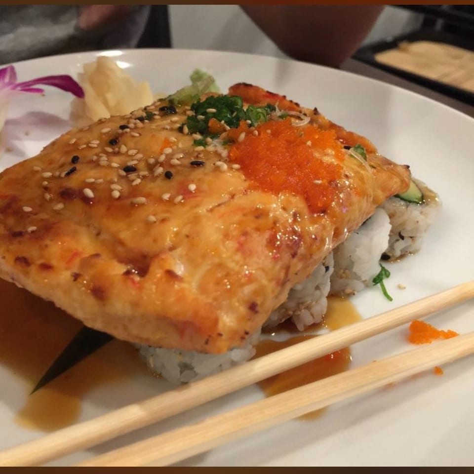 Grilled salmon sushi roll topped with orange roe, scallions, and sesame seeds on a white plate with chopsticks.
