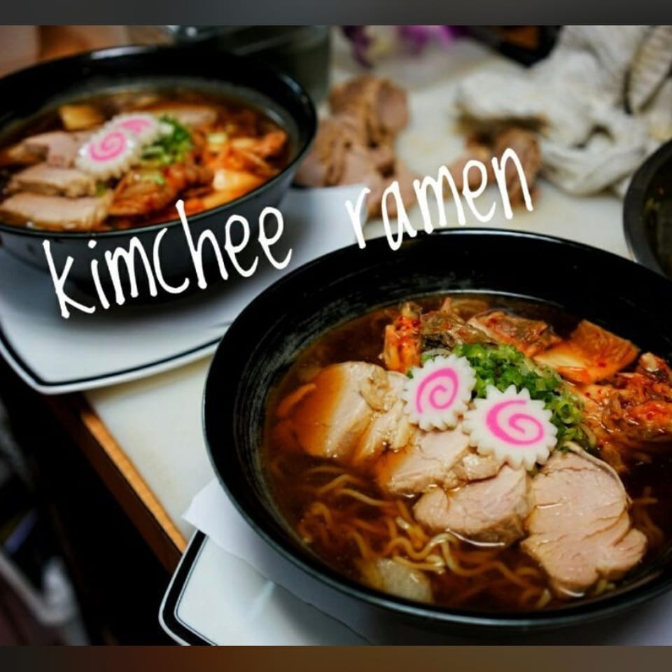 Two bowls of ramen topped with sliced pork, narutomaki, green onions, and kimchi sit on a restaurant counter.