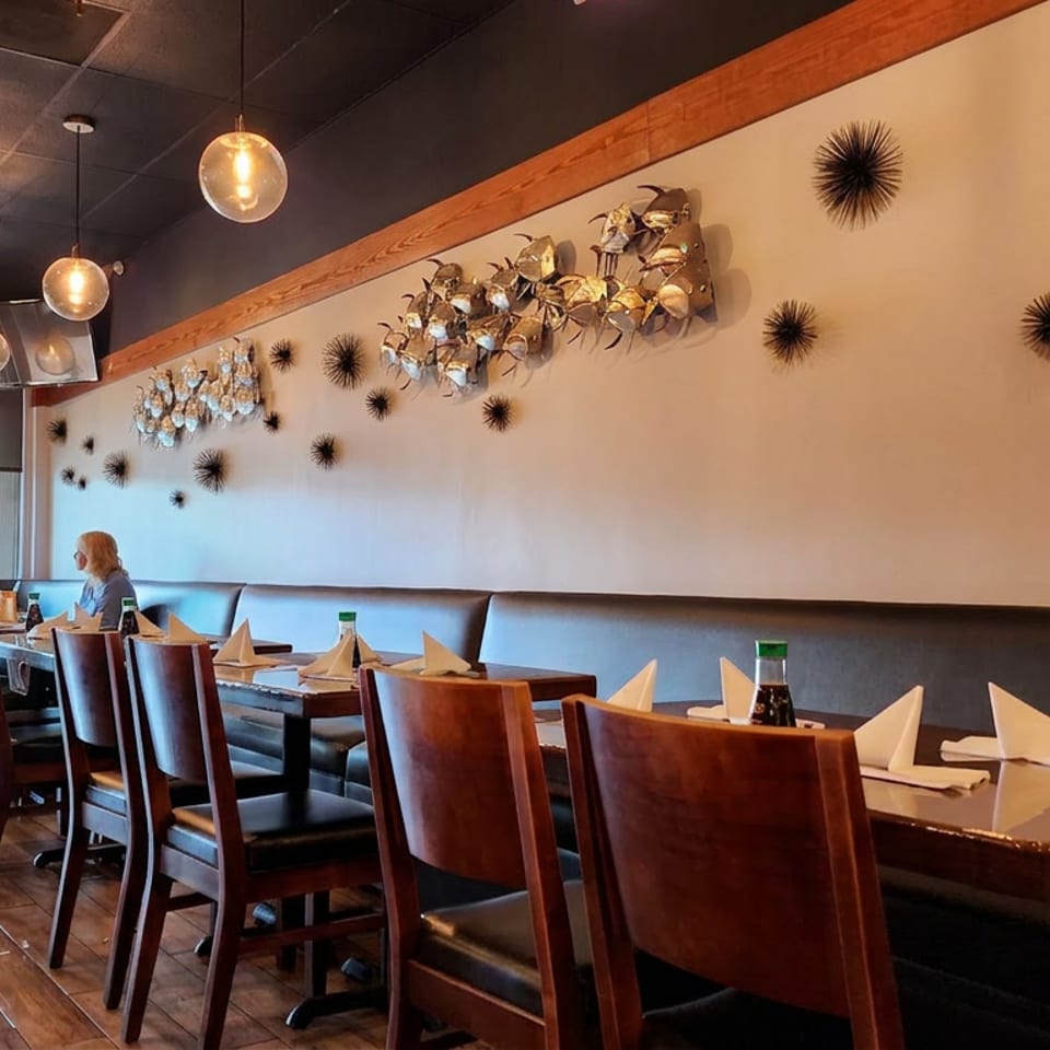 Warmly lit restaurant dining room with set tables, leather chairs, decorative wall art, pendant lights, and two diners by the window.