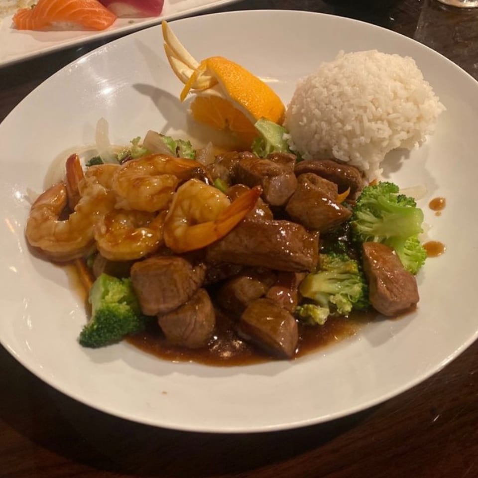 Plate of glazed shrimp, beef, broccoli, white rice, and orange garnish on a restaurant table.