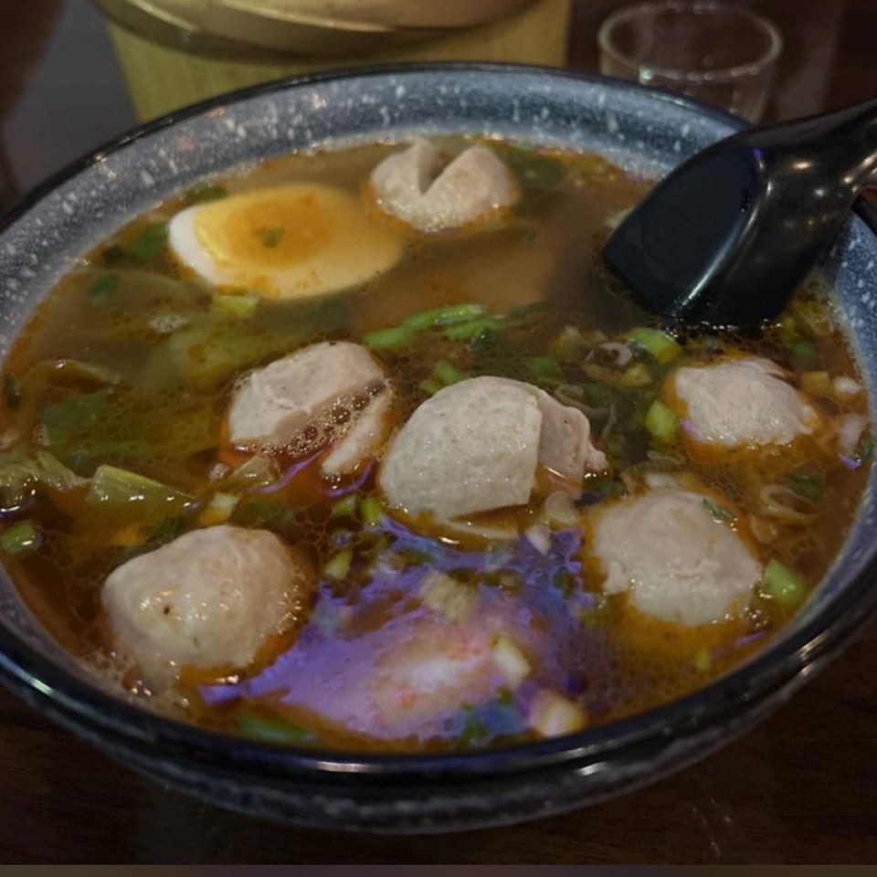 Bowl of clear broth soup with meatballs, sliced vegetables, herbs, and a black soup spoon.