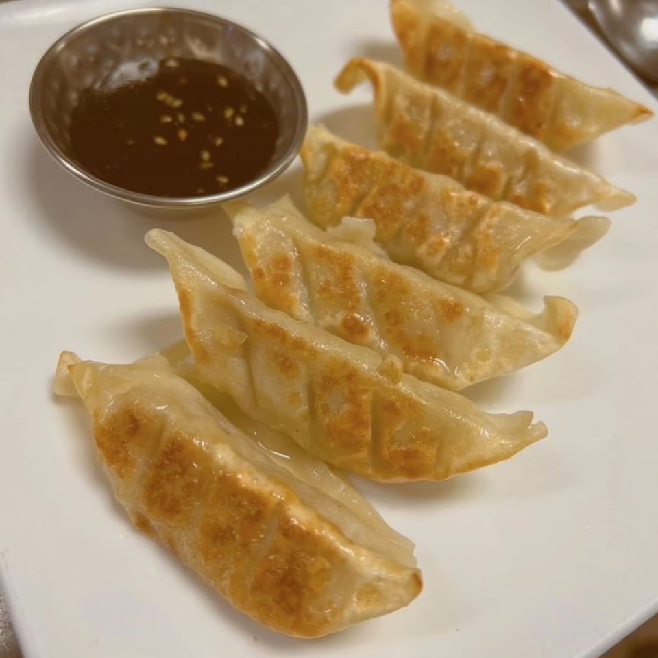 Six golden pan-fried dumplings arranged on a white plate with a small bowl of dark dipping sauce.