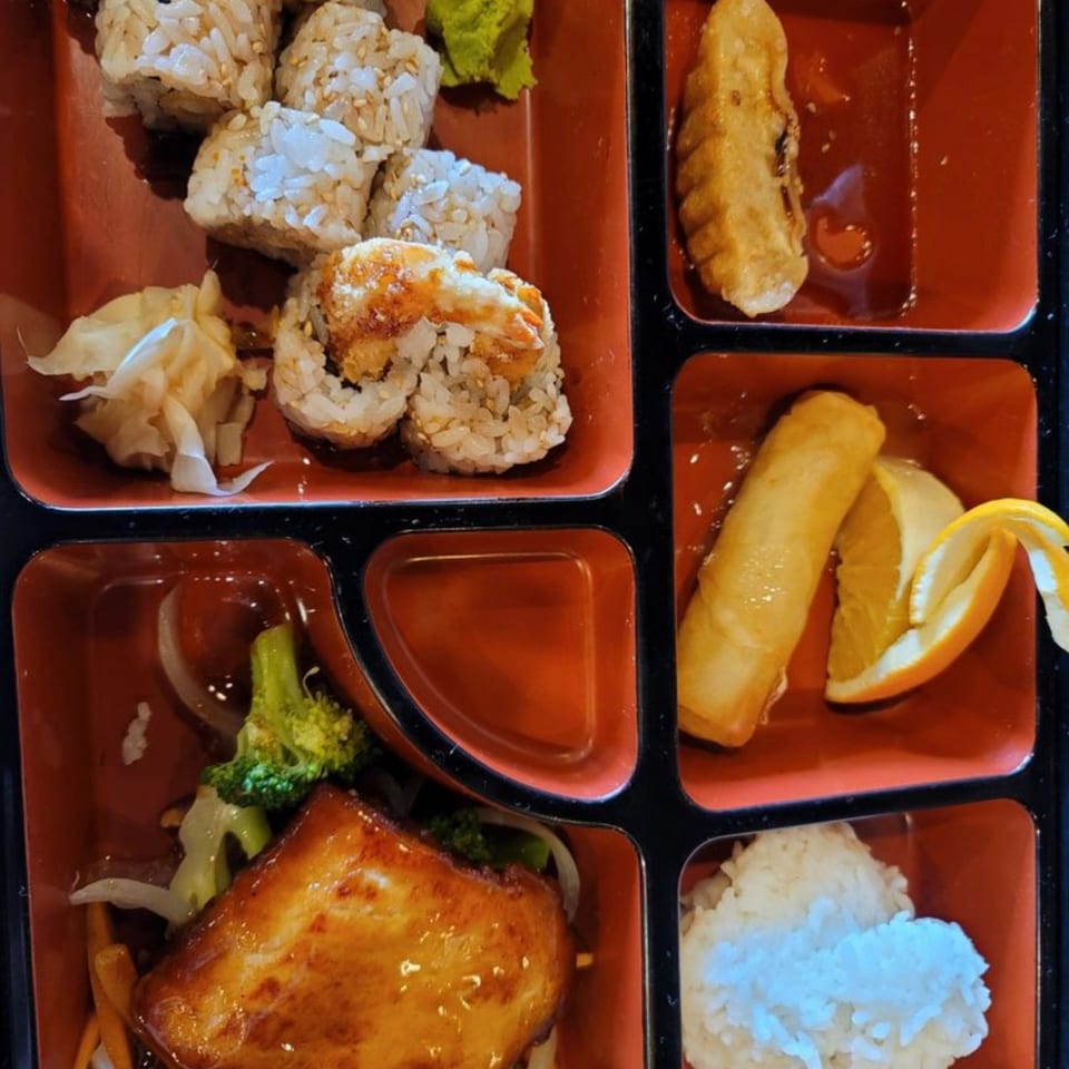 Japanese bento assortment with sushi rolls, grilled chicken, rice, spring rolls, vegetables, orange slices, wasabi, and pickled ginger.