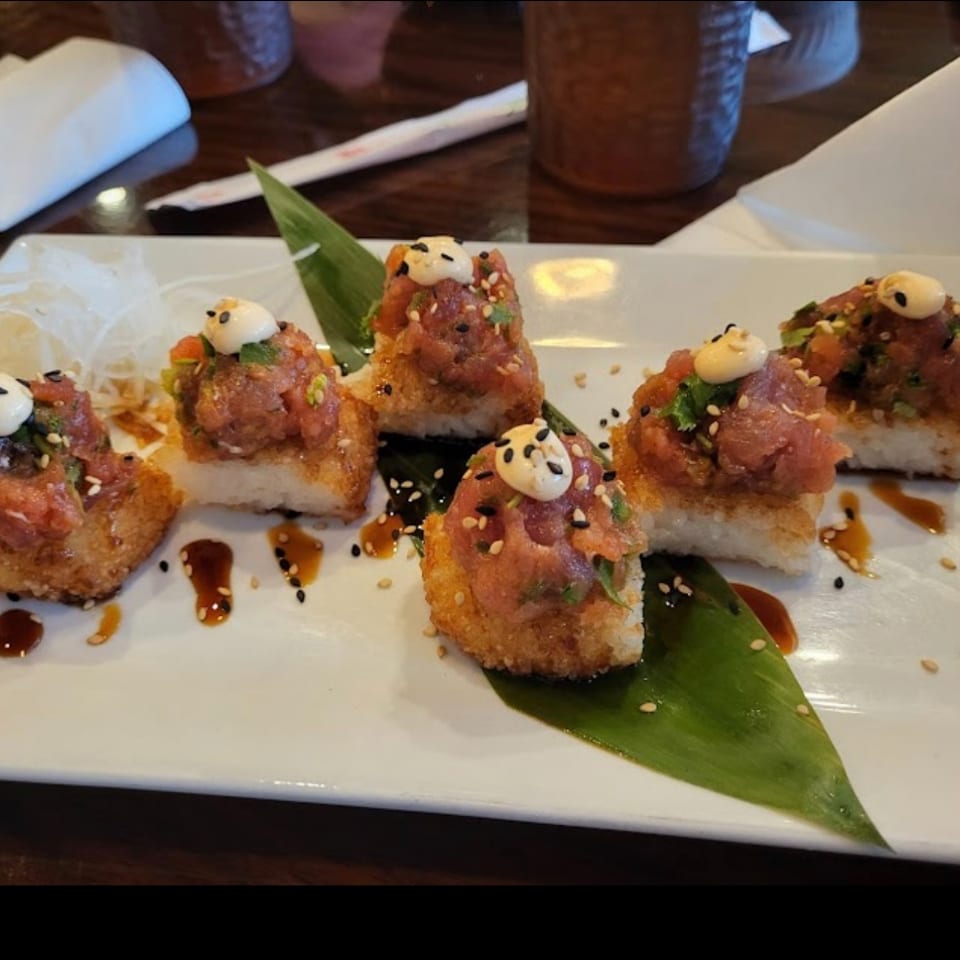 Five crispy sushi bites topped with spicy tuna, sesame seeds, and creamy sauce on a white platter.