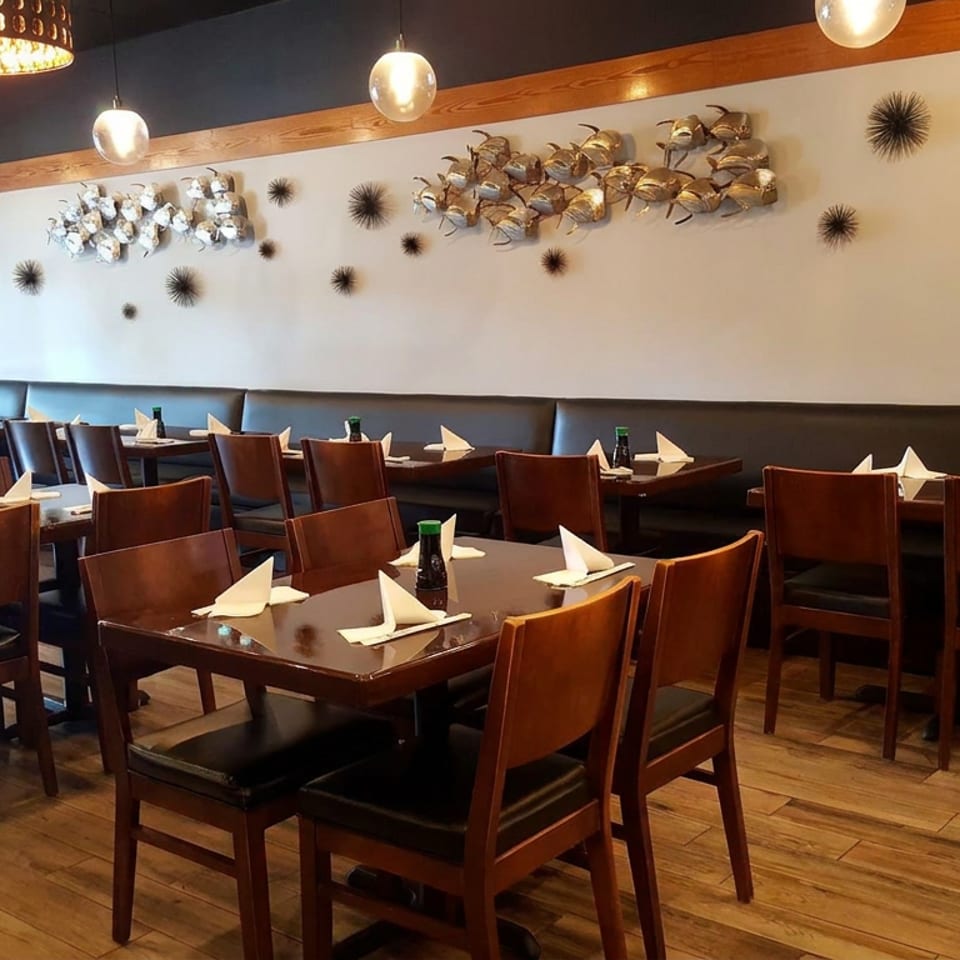 Warmly lit restaurant dining room with wooden tables, leather seating, folded napkins, decorative wall art, and two people near the entrance.