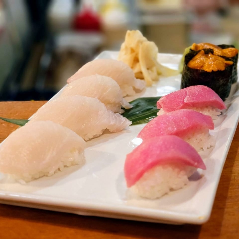 Assorted sushi nigiri and seaweed-wrapped roe sushi arranged on a white rectangular plate atop a wooden restaurant counter.