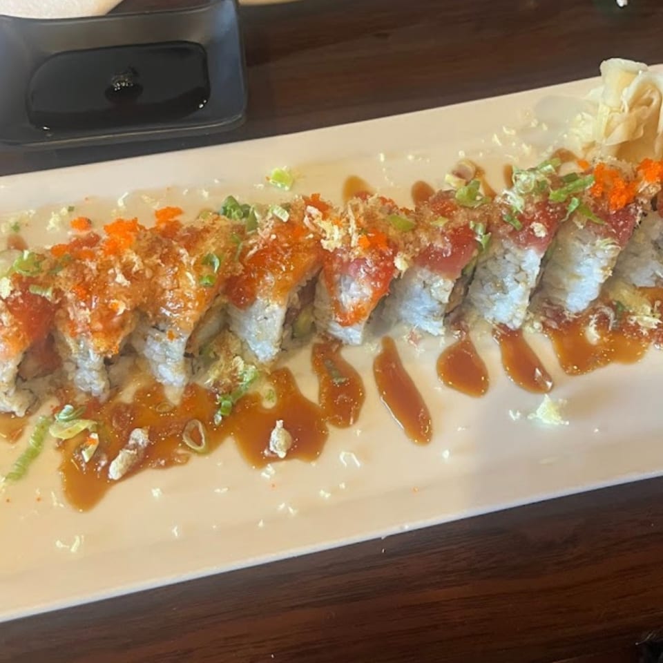 Long platter of sushi rolls topped with orange roe, green onions, crispy flakes, and drizzled sauce, accompanied by pickled ginger.