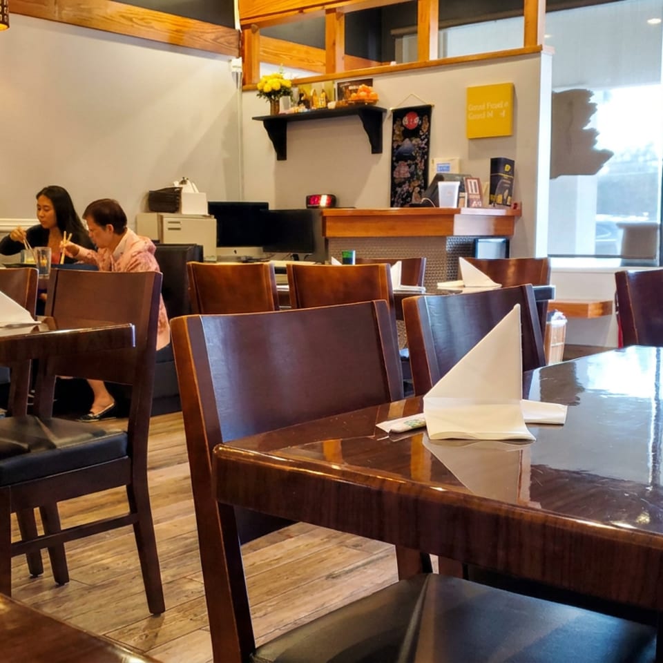 Warmly lit restaurant dining room with polished wooden tables, folded white napkins, dark chairs, and two diners near the counter.