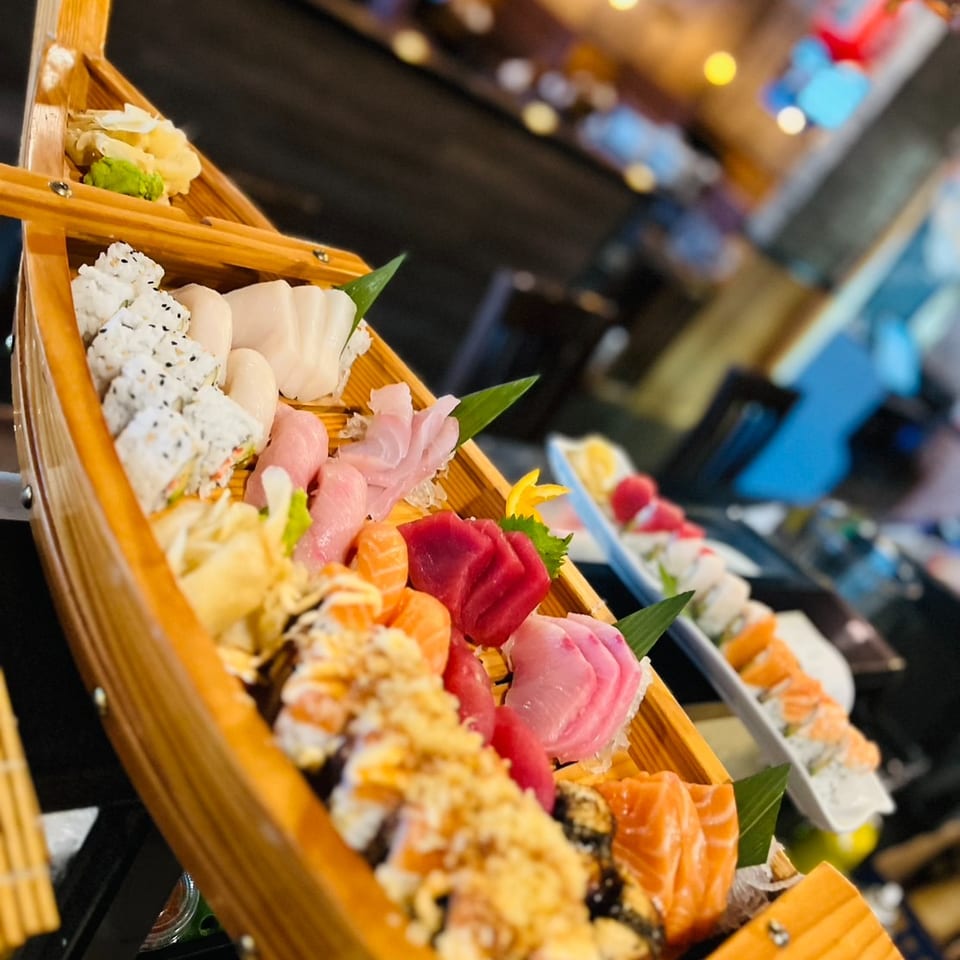 Wooden boat platter filled with sushi rolls, salmon, tuna, shrimp, and sliced sashimi at a colorful restaurant buffet.